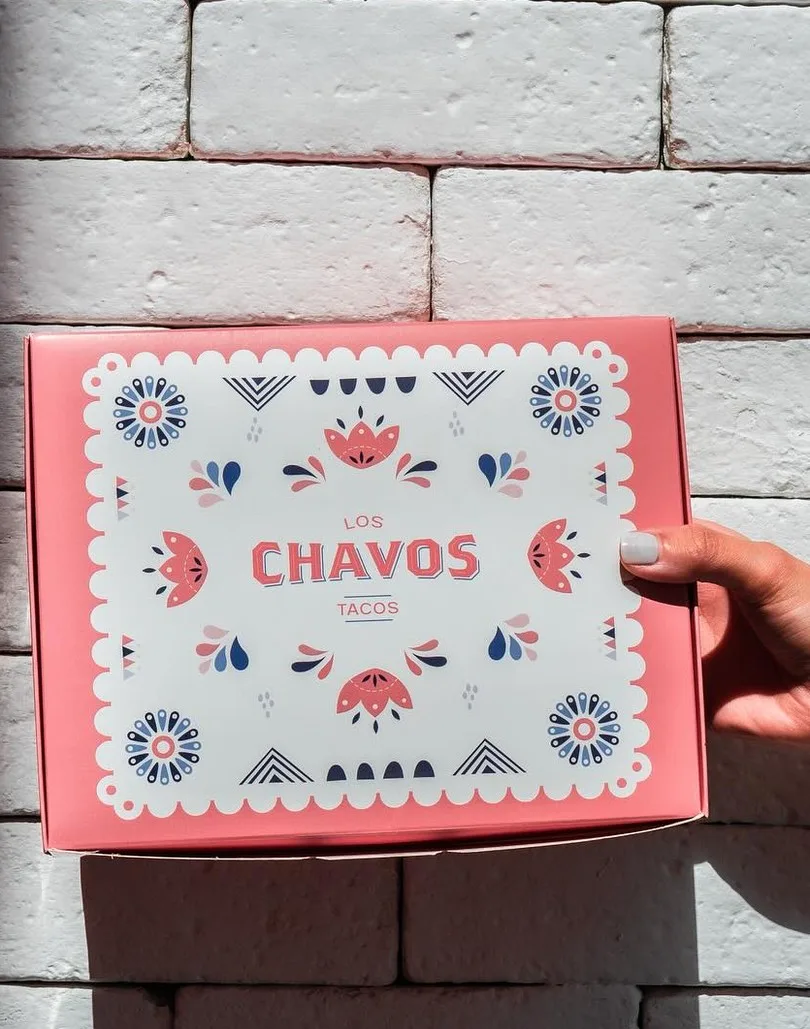 Caja rosa con marco de papel picado y el logotipo, en una mano, frente a un muro de ladrillo blanco.