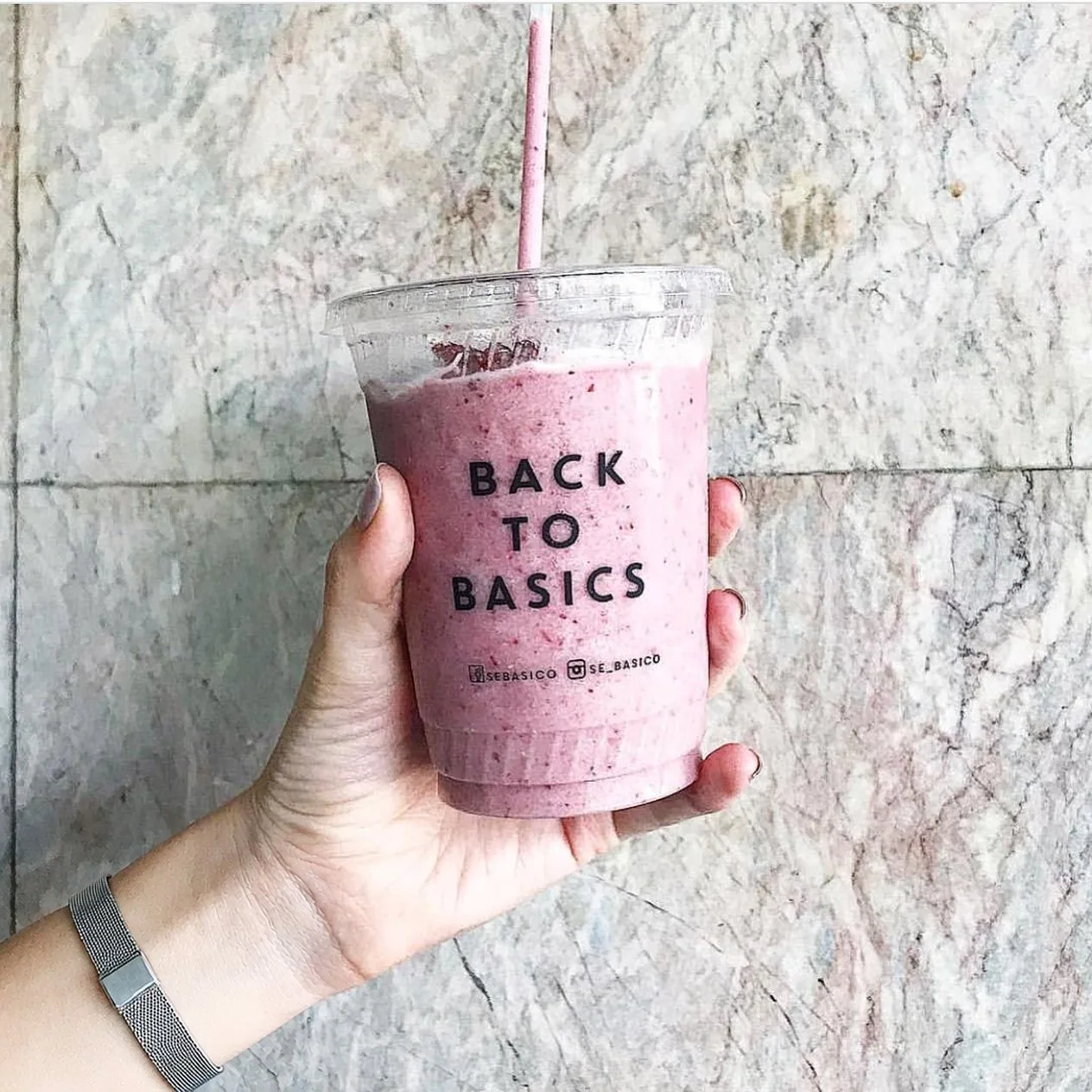 Mano que sostiene un smoothie rosa en vaso transparente con «Back to Basics» impreso.